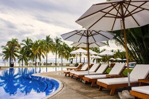 A poolside area with lounge chairs and umbrellas, palm trees, and a clear sky.