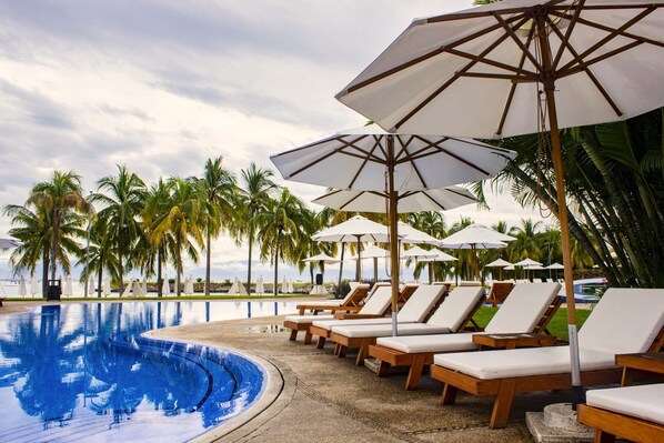 Zona junto a la piscina con sillones de descanso y sombrillas, palmeras y cielo despejado.