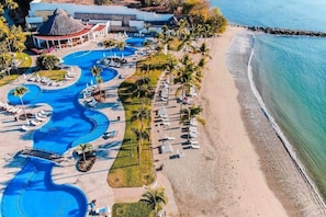 Ein Luftbild eines Resorts mit mehreren Schwimmbecken, einem Strand und einem großen Gebäude mit einem Reetdach.