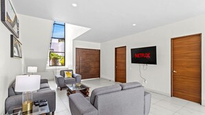 A modern living room with grey sofas, a coffee table, a TV mounted on the wall, and a large window.