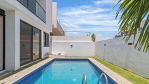 Un área de piscina moderna con una piscina rectangular, una escalera metálica y una pared blanca con un ventanal.