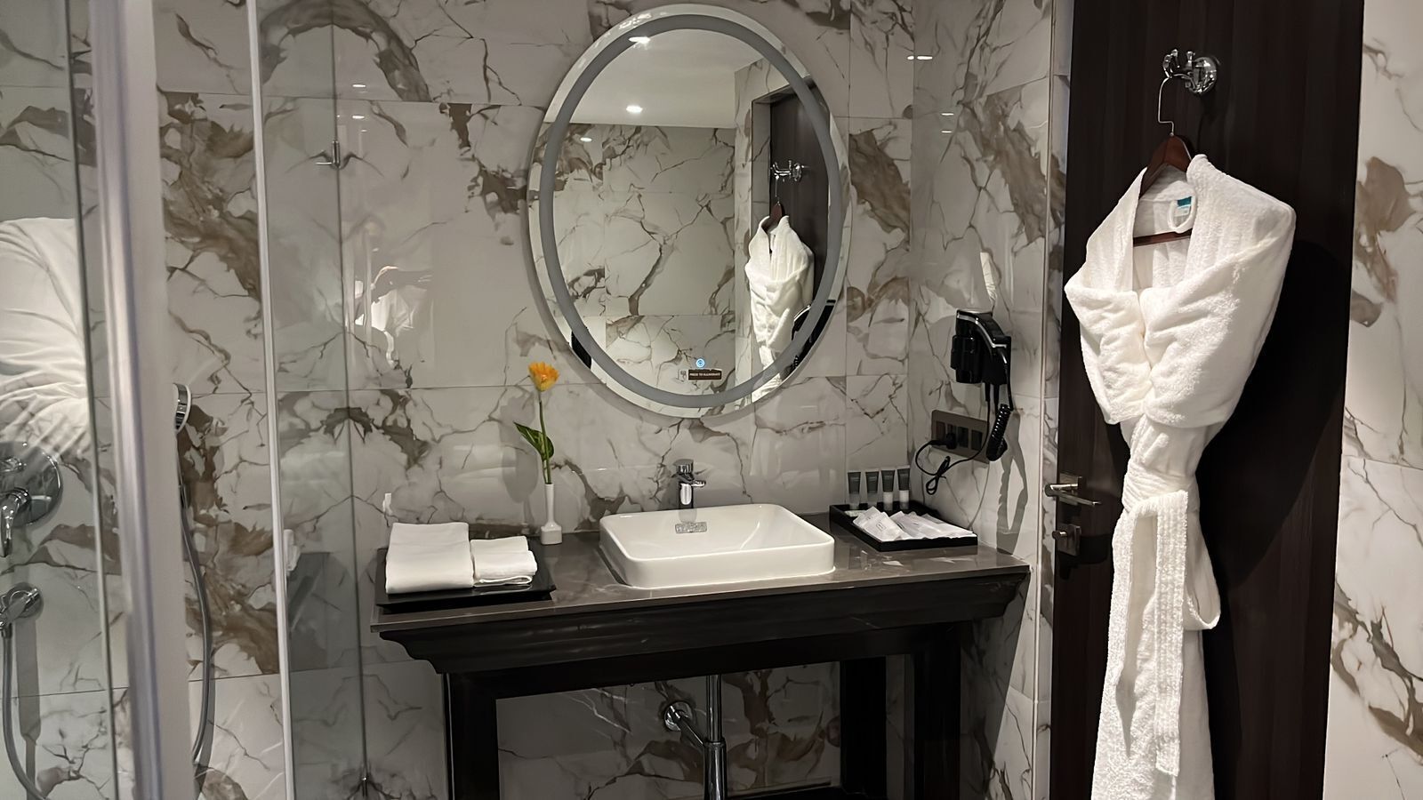 Ein Badezimmer mit Marmor-Spritzschutz, einem runden Spiegel, einem weißen Waschbecken, einem Handtuchhalter und einem Bademantel, der an der Wand hängt.