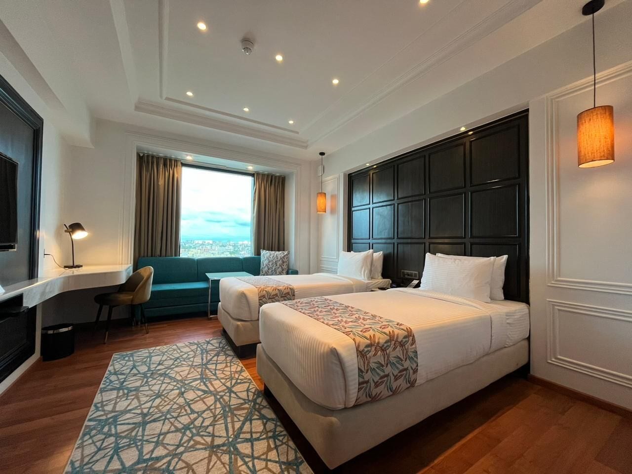 Habitación de hotel con dos camas, un escritorio, una silla, un ventanal con vistas y una alfombra estampada.