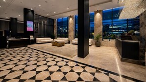En moderne lobby med et skakternet gulv, sofaer, en receptionsdisk og store vinduer med udsigt over byen.