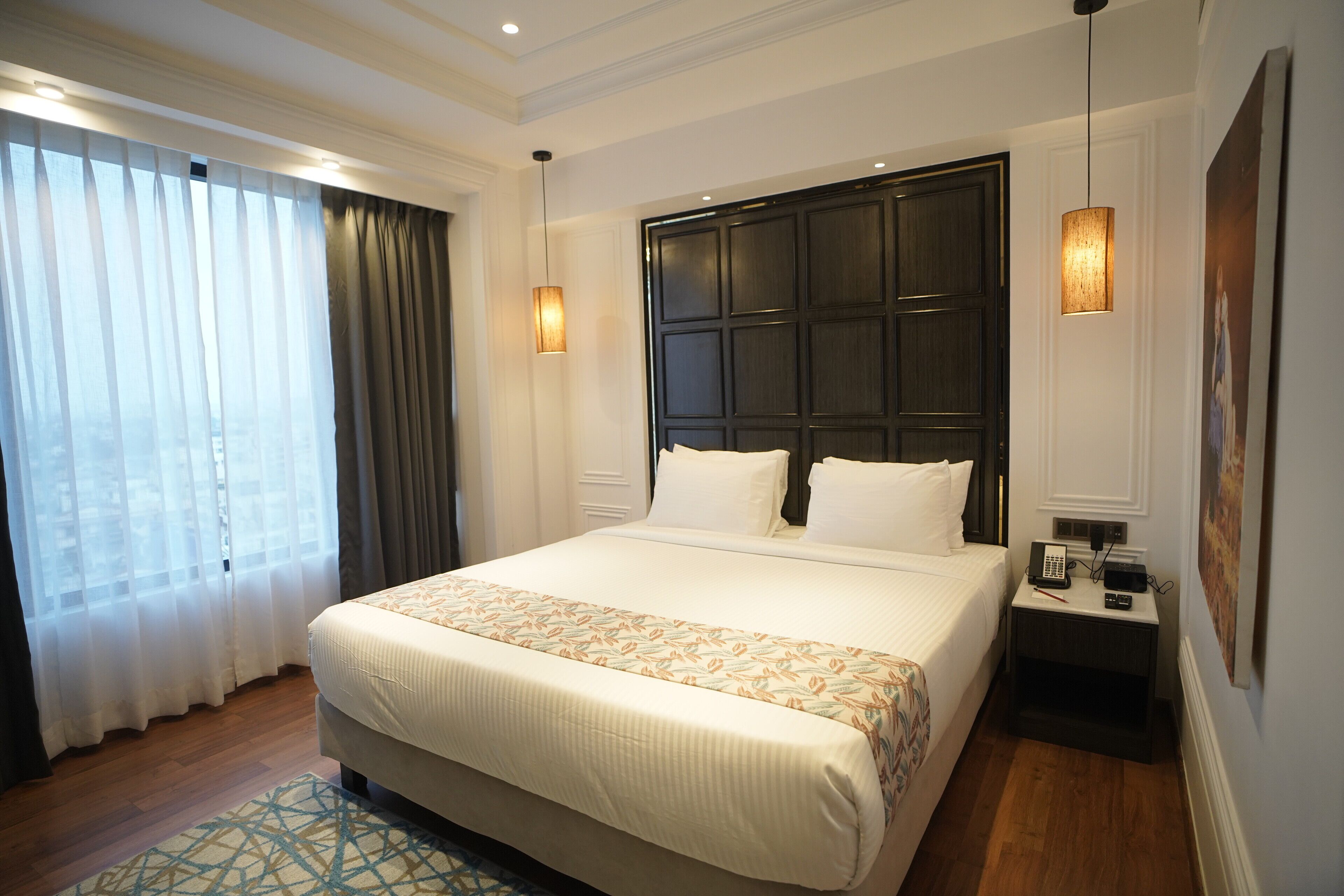A hotel room with a large bed, a nightstand with a phone and remote, a window with curtains, and a wall-mounted light fixture.