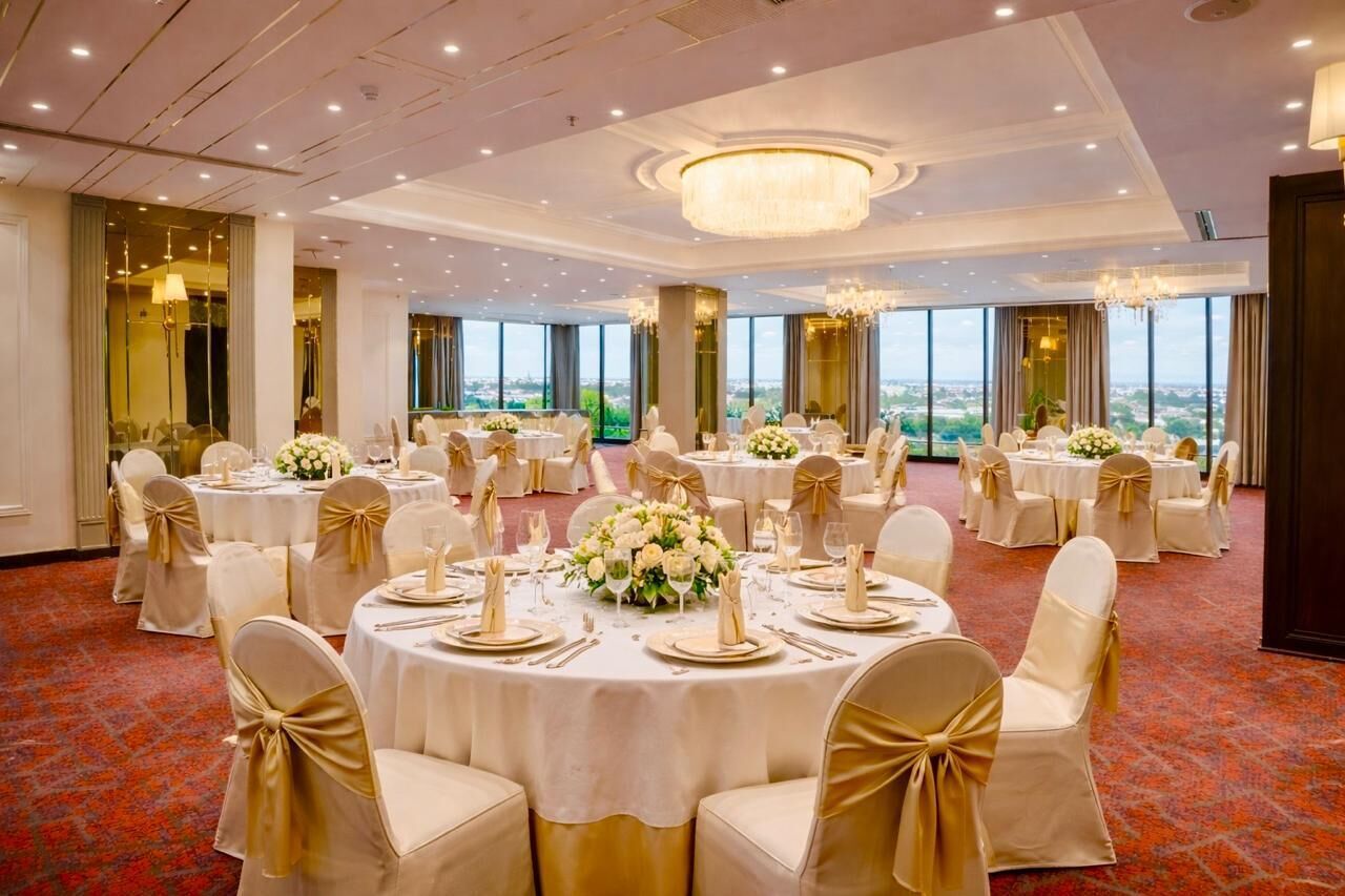Un salón de banquetes con mesas redondas, manteles blancos y cintas doradas en las sillas.