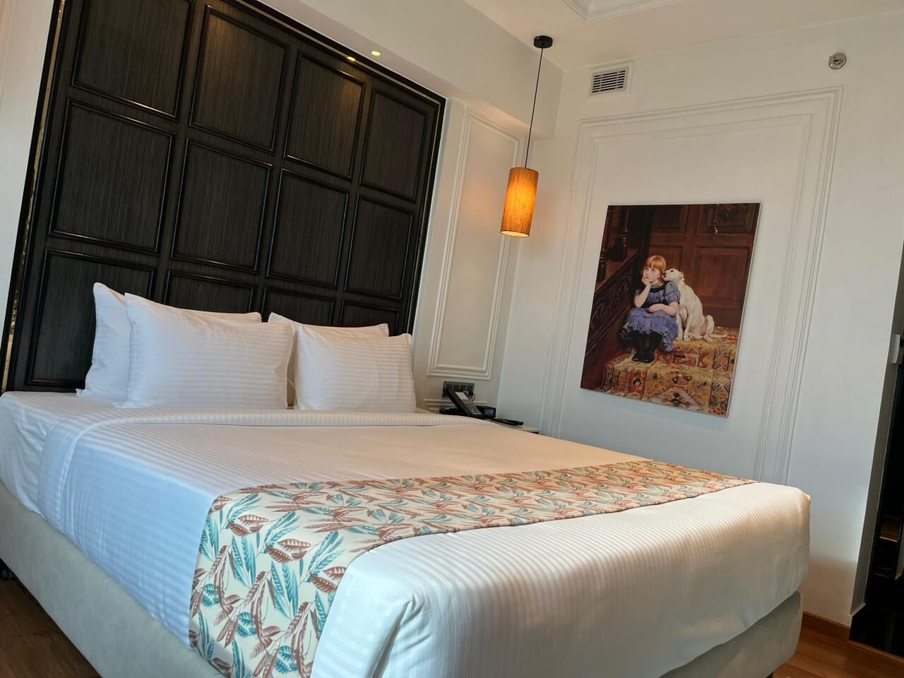 Una cama bien hecha con sábanas blancas y un plaid estampado floral, cabecera de madera oscura, una lámpara colgante y un cuadro en la pared.