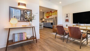 A modern living room with a dining area, a kitchen in the background, and a flat-screen TV mounted on the wall.