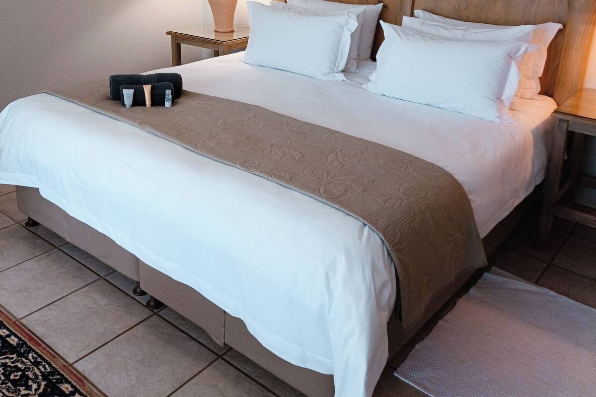 Een opgemaakt bed met houten hoofdbord, een nachtkastje met lamp en een ingelijst beeld aan de muur.