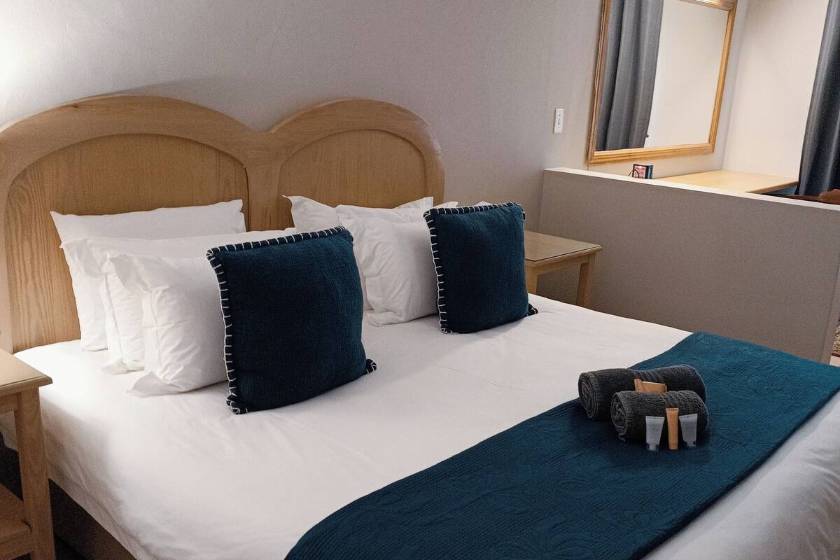 Een net opgemaakt bed met witte en donkerblauwe kussens, een blauwgroene deken en een klein nachtkastje met een lamp.