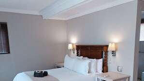 A neatly arranged hotel room with a large bed, two bedside tables, a small nightstand, a wall-mounted lamp, and a window with blinds. - Pecan Farm Guesthouse (Louis Trichardt)
