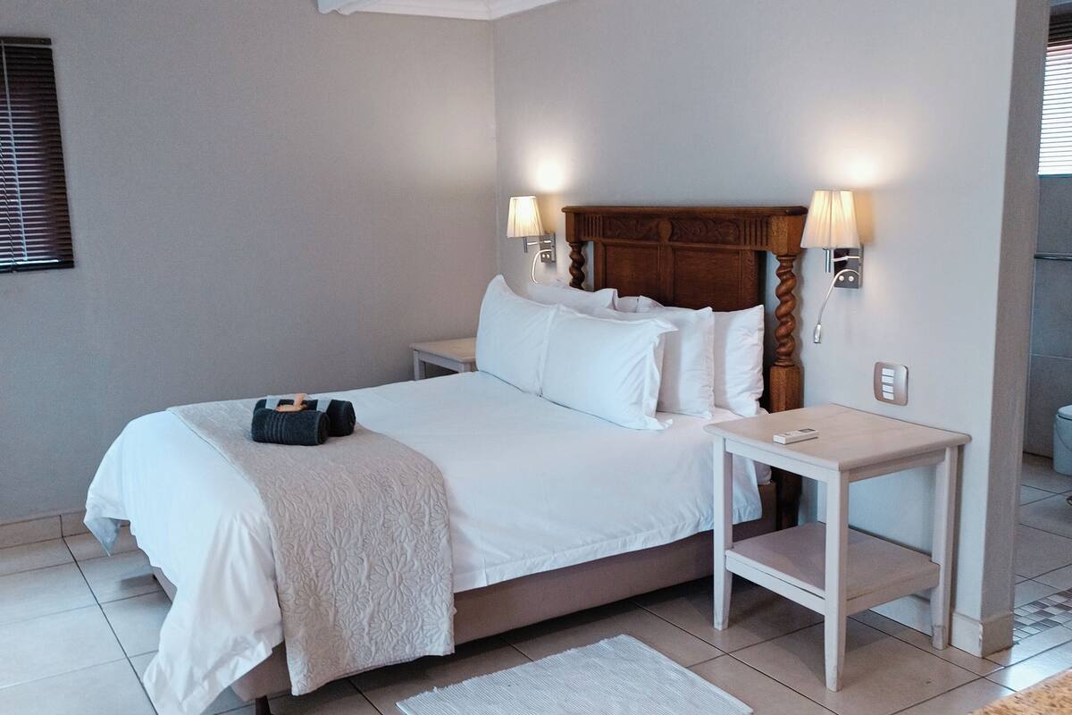 Een keurig opgeruimde hotelkamer met een groot bed, twee nachtkastjes, een klein bijzettafeltje, een wandlamp en een raam met zonwering.