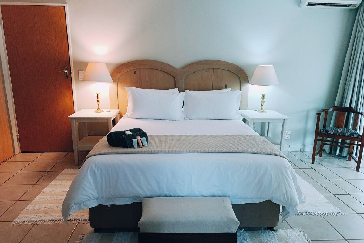 Een opgemaakt bed met witte lakens, twee nachtkastjes met lampen, een houten hoofdbord, een stoel en een deur.