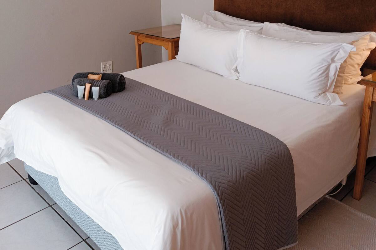 Een opgemaakt bed met witte en grijze beddengoed, een houten nachtkastje en een bruin hoofdbord.