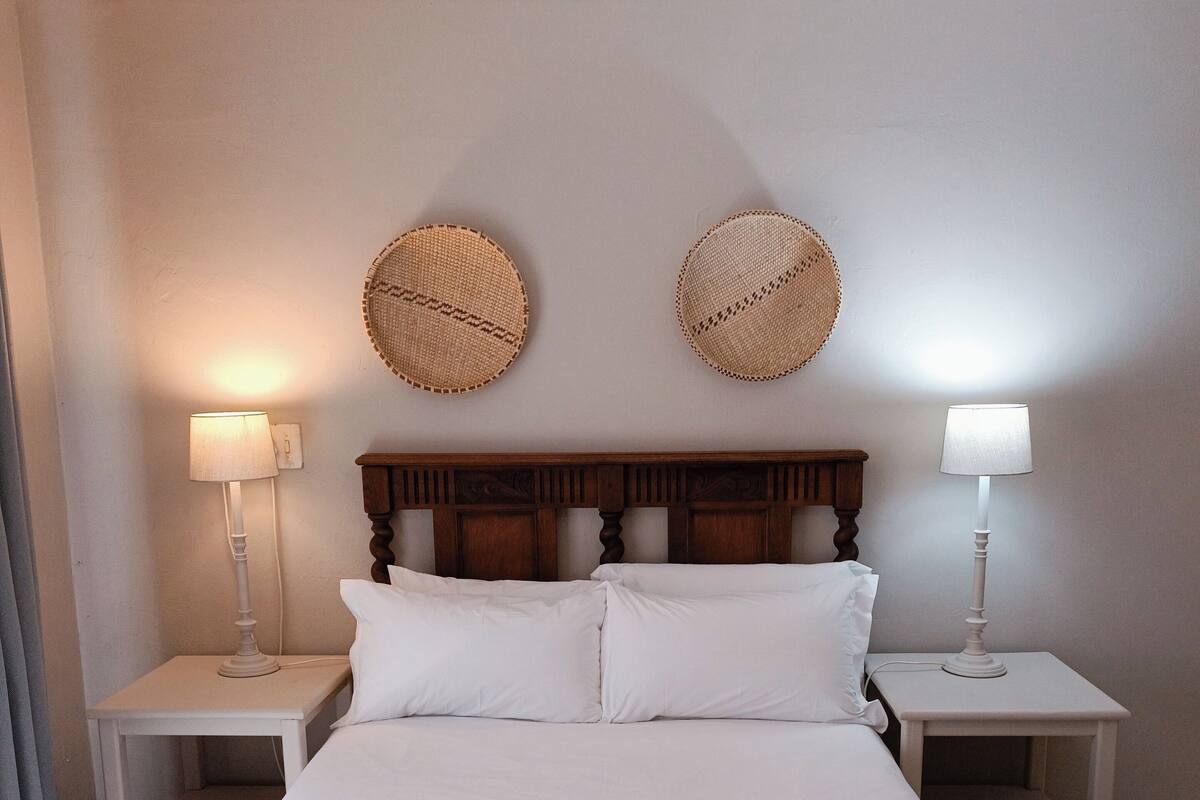 Een keurig opgemaakt bed met witte lakens, twee nachtkastjes met lampen, en twee geweven wanddecoraties.