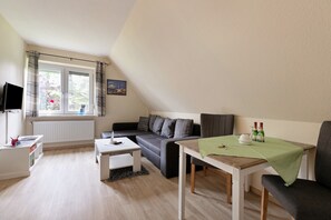 A modern living room with a sofa, a coffee table, a small table with a green tablecloth, and a television on a stand.