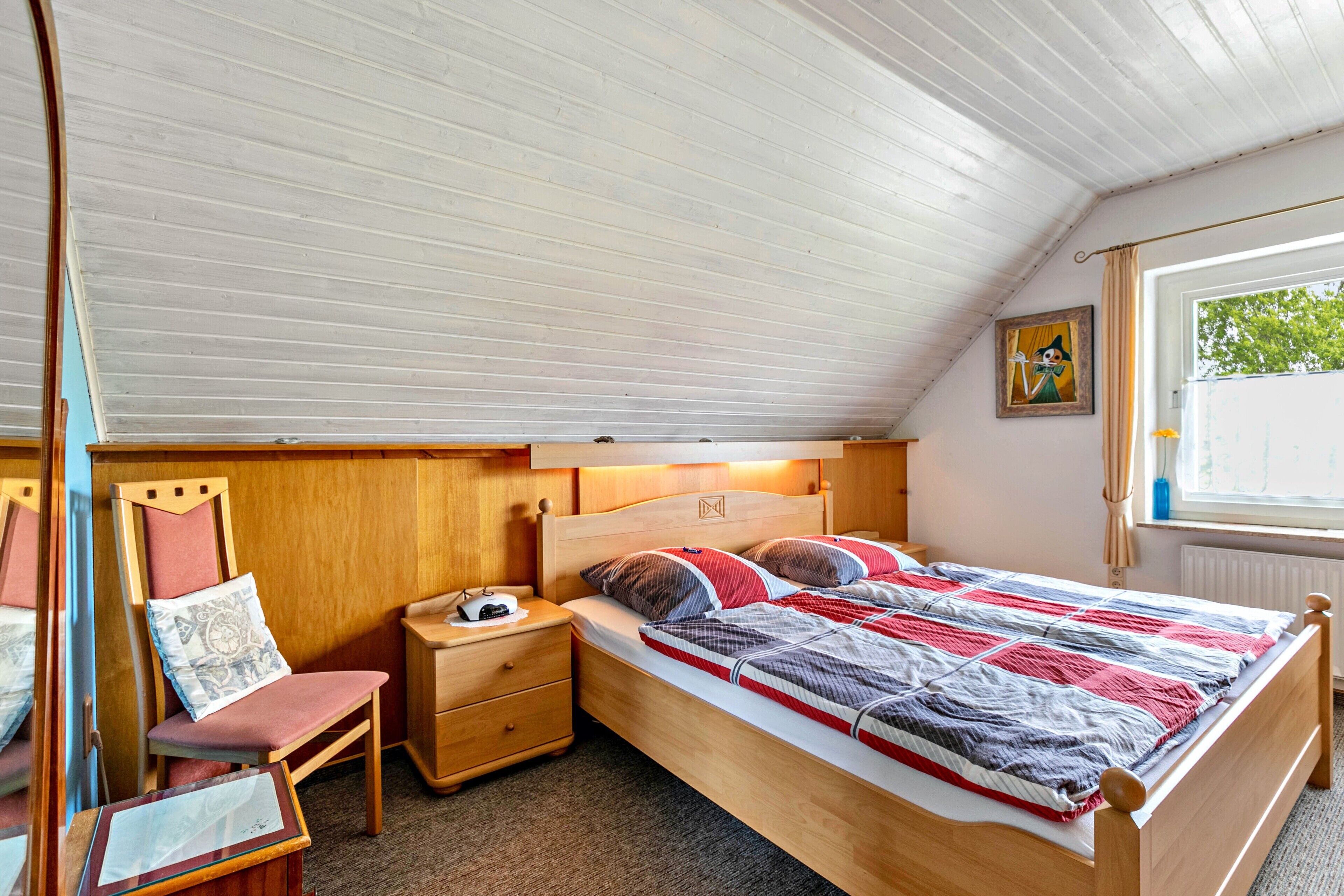 A single bed with a wooden headboard, a bedside table, a chair, and a window with a view of greenery.