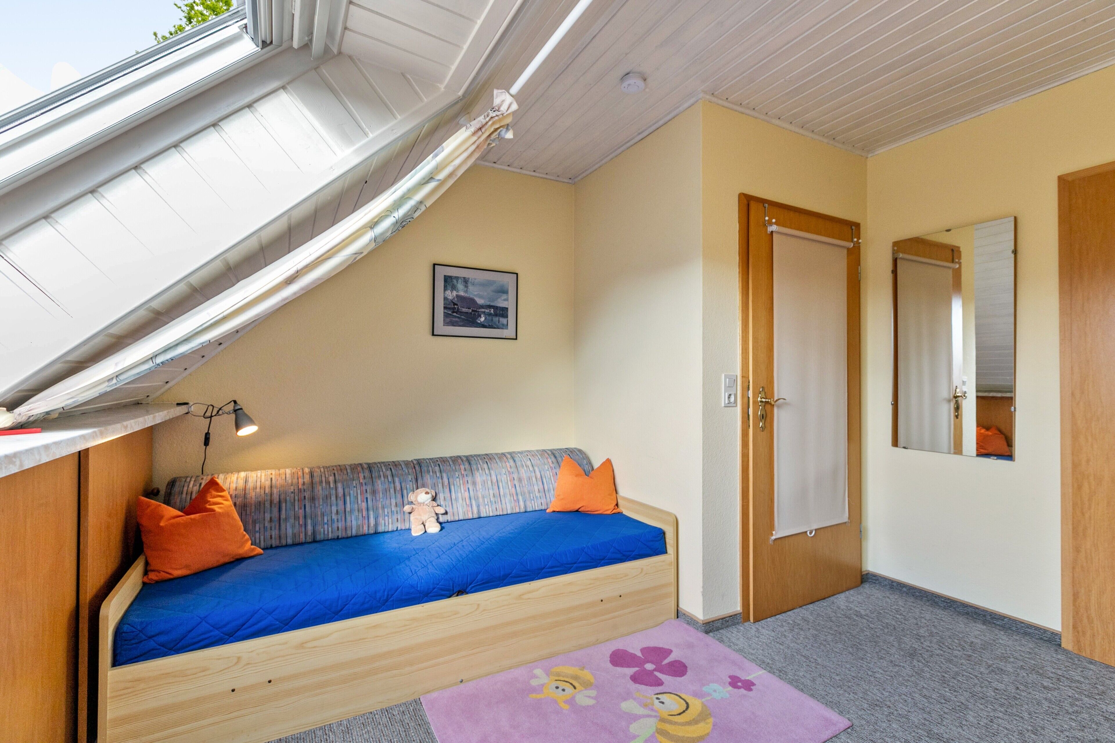 A room with a bunk bed, a small table, a lamp, a door, a mirror, and a window with curtains.