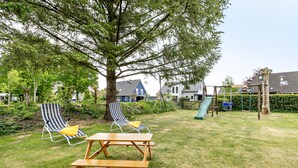 Een achtertuin met een picknicktafel, twee gestreepte lounge fauteuils, een boom en een speeltuin met een glijbaan.