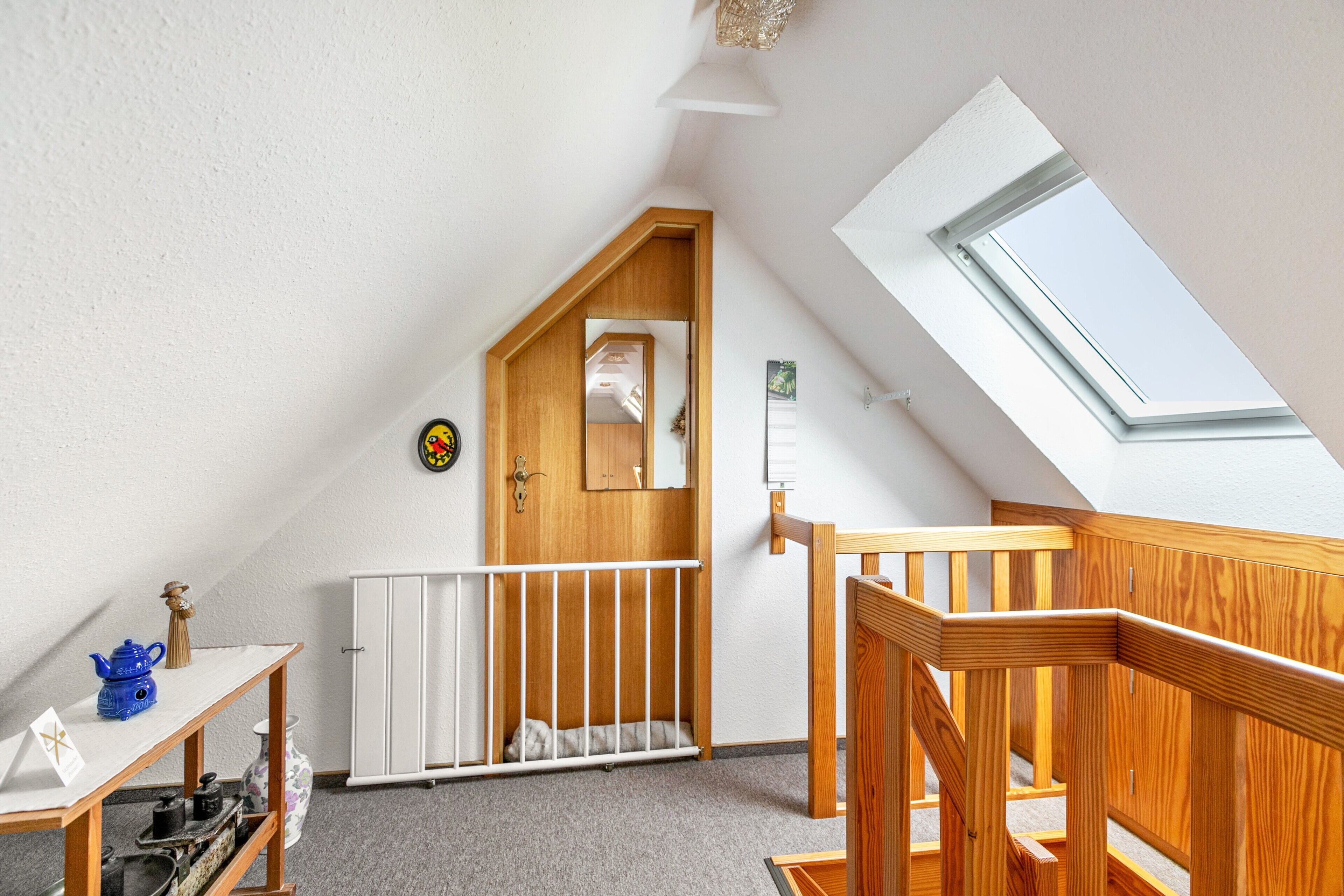 Ein Zimmer mit schrägverdeckter Decke, einer Holztür, einem weißen Heizkörper, einem weißen Kinderbett, einer Holztreppe und einem Oberlicht.