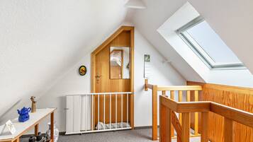 Ein Zimmer mit schrägverdeckter Decke, einer Holztür, einem weißen Heizkörper, einem weißen Kinderbett, einer Holztreppe und einem Oberlicht.