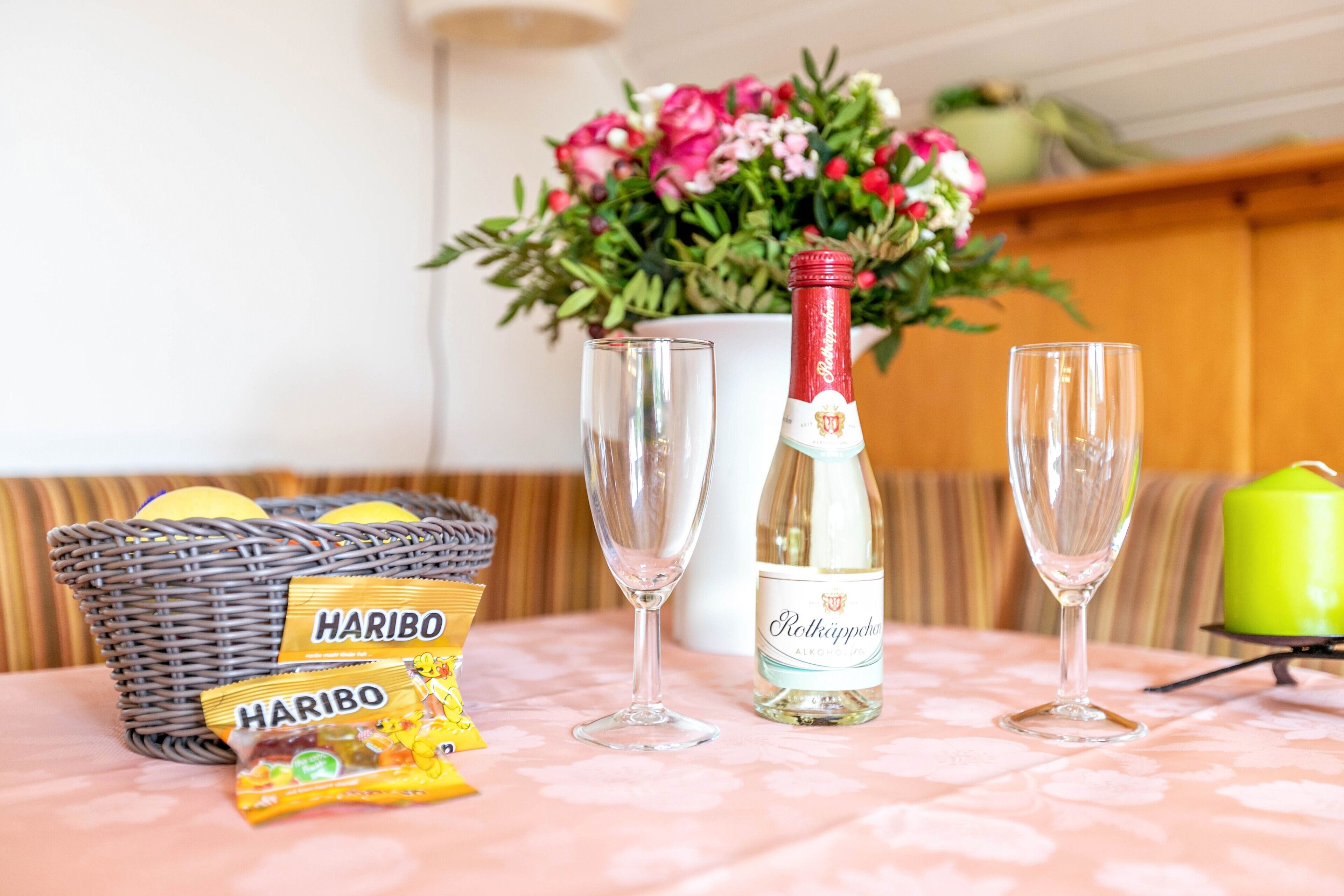 A table with a bottle of Röllicsperch, two glasses, a basket of Haribo candies, and a vase of flowers.
