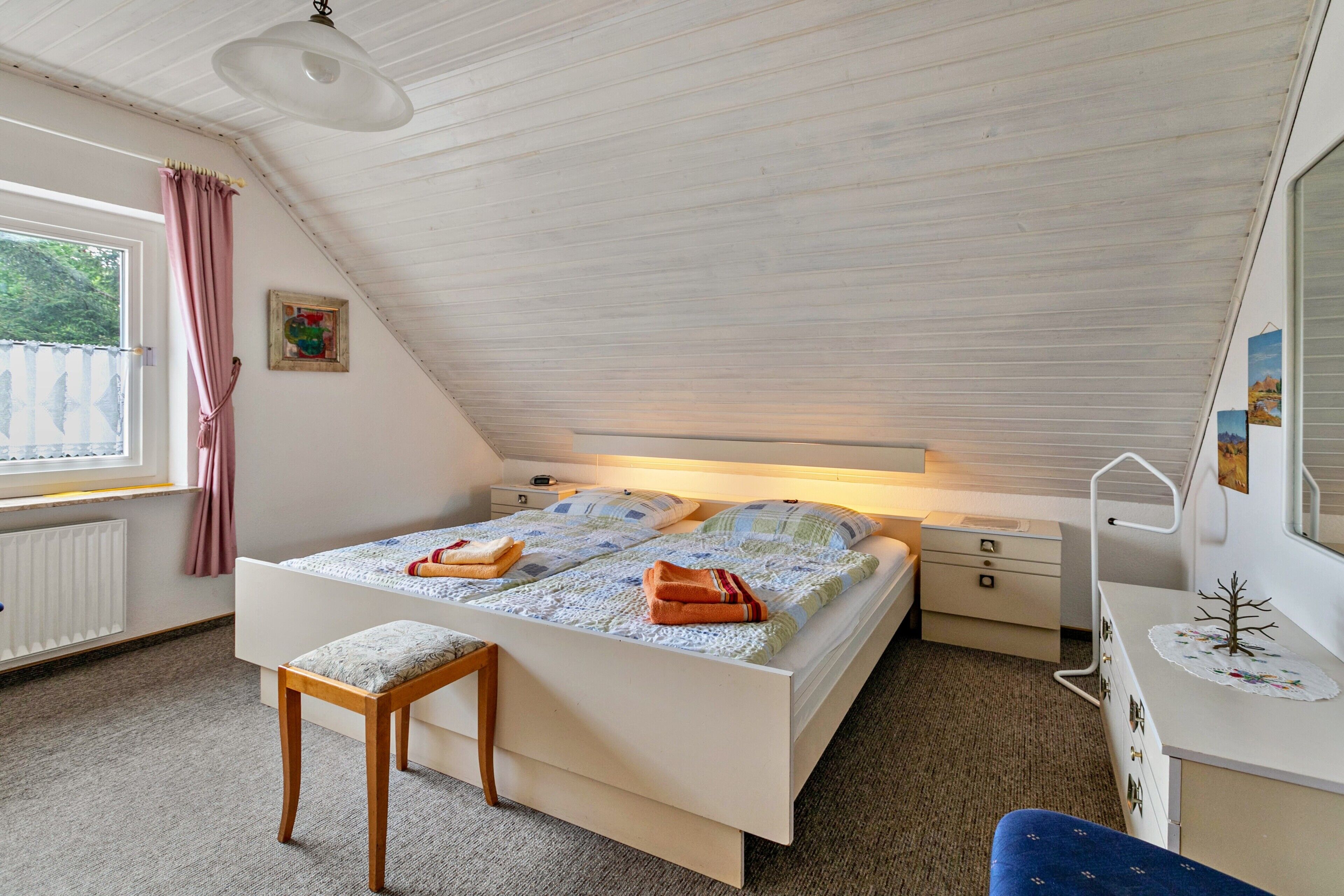 A bedroom with a sloped ceiling, a bed with a quilt, a bedside table, a chair, a dresser, and a window with curtains.