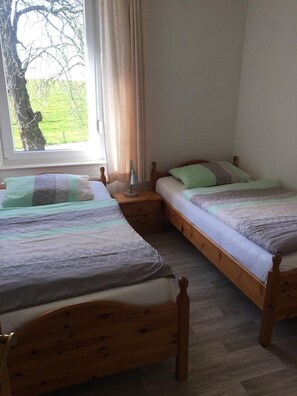 Ein Zimmer mit zwei hölzernen Betten, einem Nachttisch, einer Lampe und einem Fenster mit Blick auf Baum und Rasen.