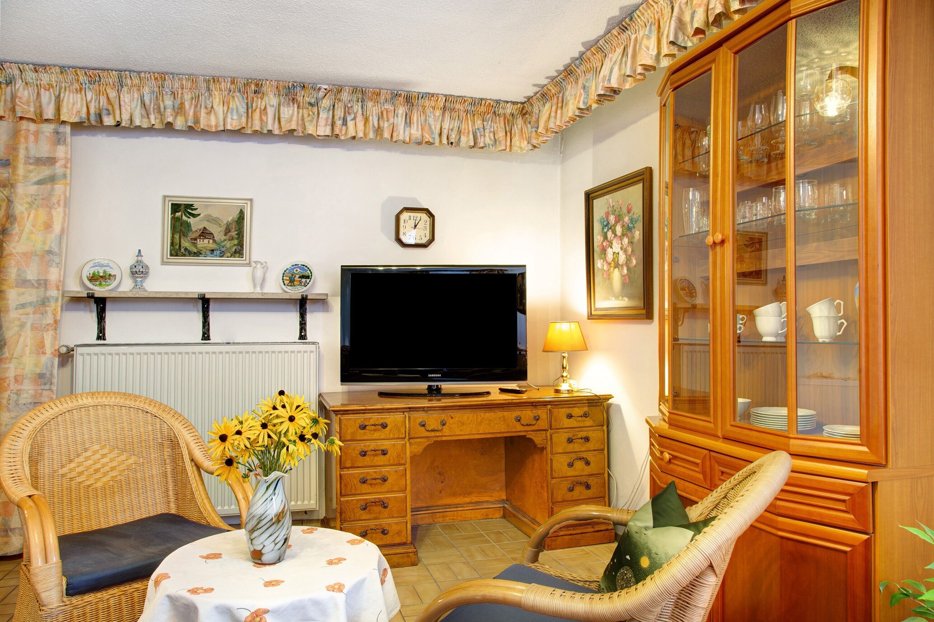 A living room with a television, wooden desk, wicker chairs, a vase with flowers, and a cabinet with glass doors.