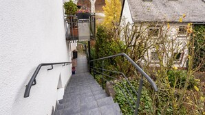 Un escalier menant à un bâtiment blanc orné d’une rambarde en métal, avec un pot de fleurs et, au loin, une vue sur un pont.