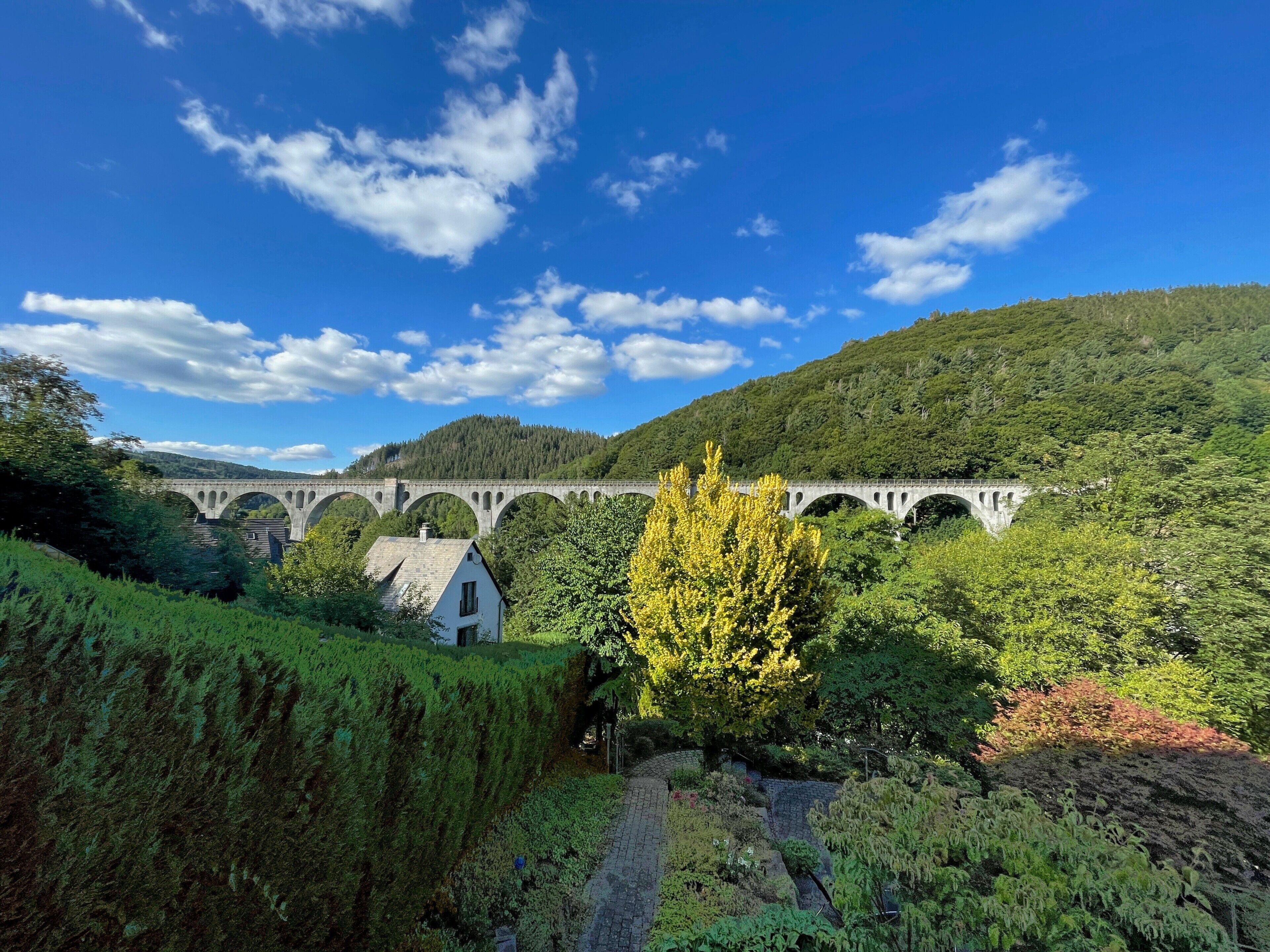 A stone bridge over a river, surrounded by lush greenery and a small white building.