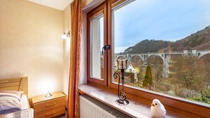 Une chambre à coucher comprenant un lit, une table de chevet, une lampe, un radiateur et une fenêtre donnant sur un pont et des arbres.
