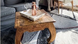Uma sala de estar com sofá cinza, mesa de centro de madeira com bandeja, luminária de chão, poltrona branca e fofa, e janela com cortinas.