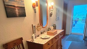 Um banheiro com cadeira de madeira, toucador com espelho e vista para o exterior através de uma porta de vidro.