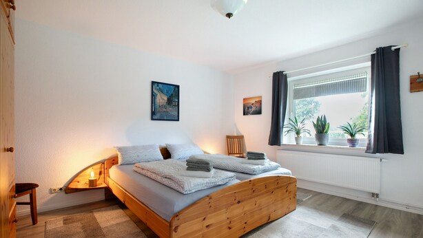 A bedroom with a wooden bed, a nightstand, a lamp, a window with curtains, and two wall-mounted pictures.