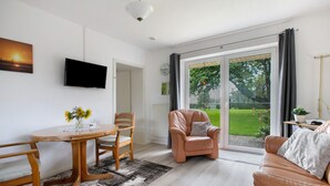 A modern living room with a sofa, a chair, a dining table with chairs, a flat-screen TV mounted on the wall, and a large sliding glass door leading to a garden.