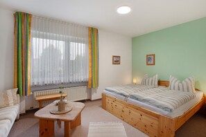 A bedroom with a wooden bed, a bedside table, a window with sheer curtains, a radiator, and a small picture on the wall.