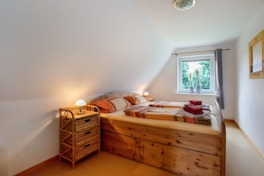 Une chambre avec un plafond en pente, un lit doté d’une tête de lit en bois, une table de chevet avec une lampe, une fenêtre donnant sur un paysage verdoyant et un cadre photo fixé au mur.