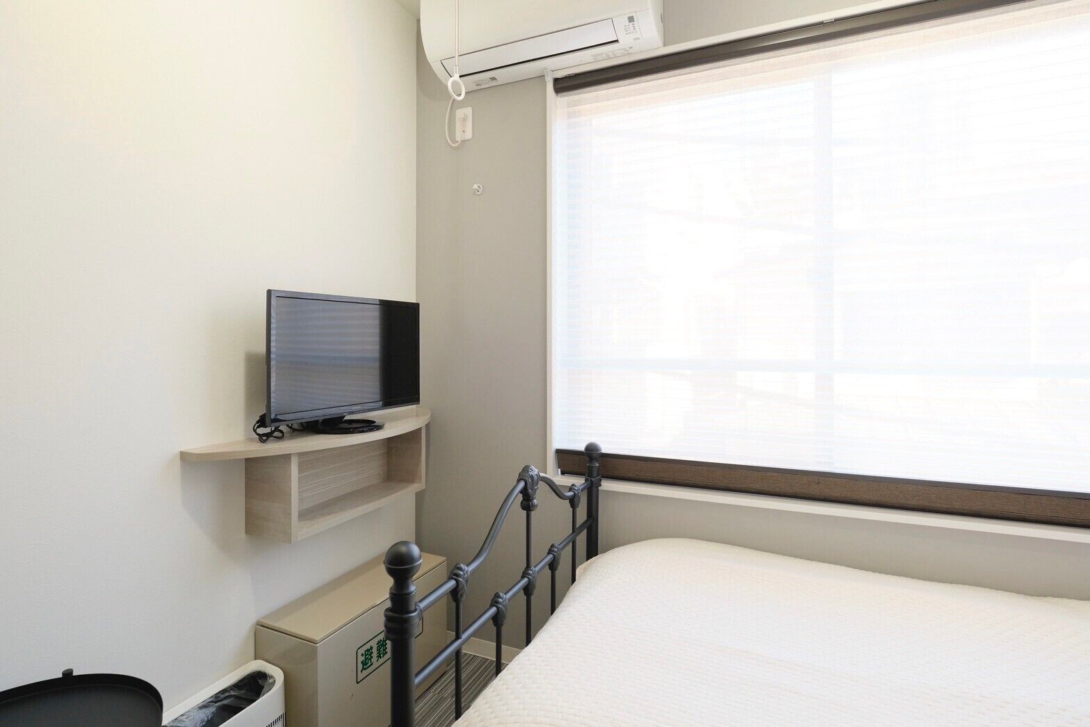 A hotel room with a bed, a television on a shelf, a window with blinds, and a radiator.