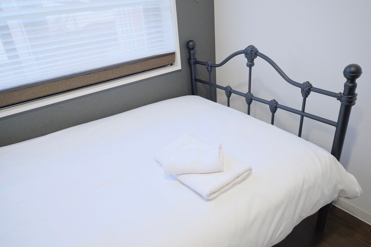 A neatly made bed with a white sheet and a folded towel, a metal headboard, and a window with blinds.