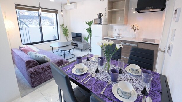 A modern living room with a purple sofa, a dining table set with plates, cups, and cutlery, and a kitchen area with a microwave and stove. - HJ PLACE TAKANAWA by Tranova (Tokyo)