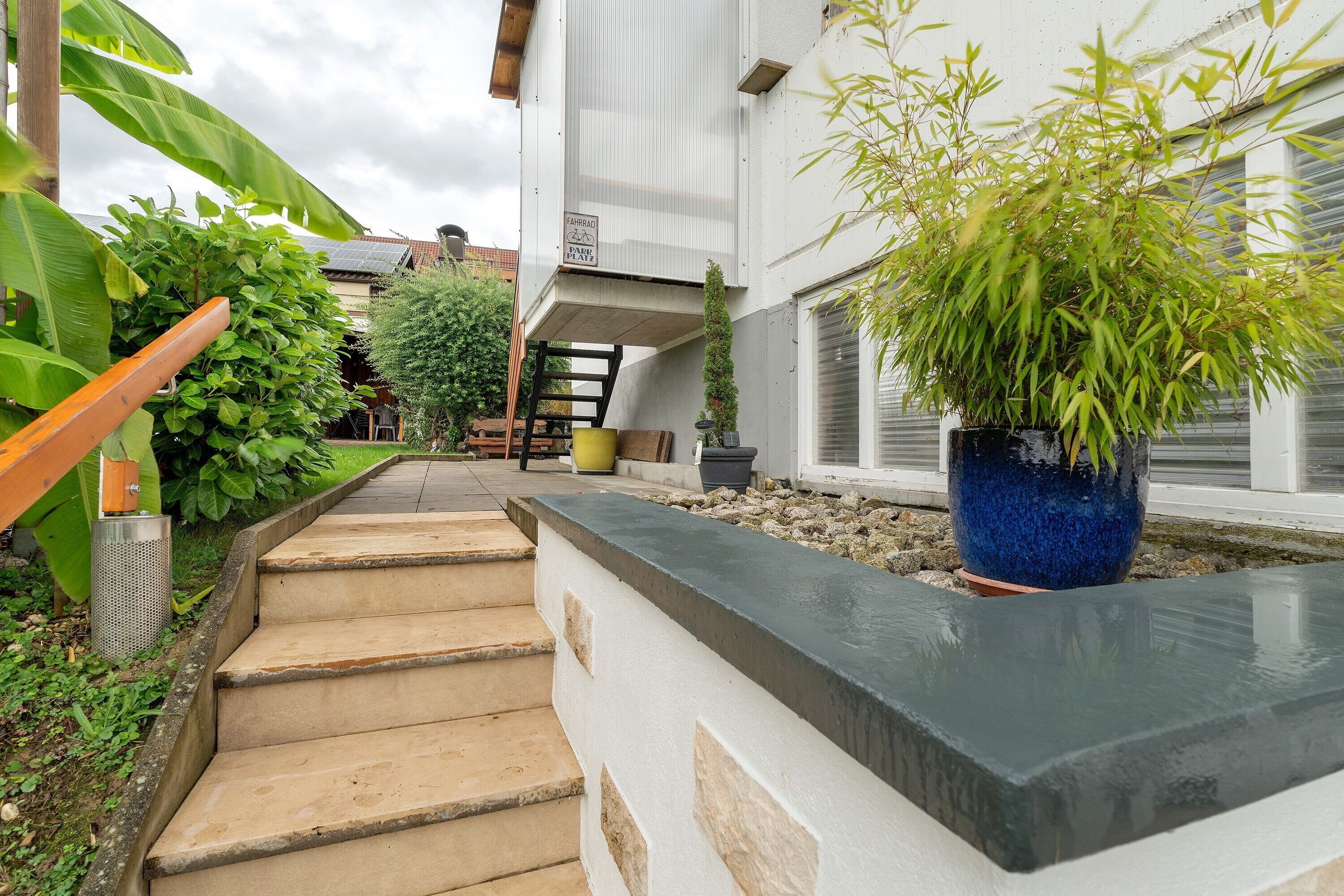 Eine Treppe führt zu einem Hauseingang, ein großer Blumentopf und ein Metallmülleimer.