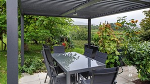 A covered patio with a glass table and chairs, surrounded by lush greenery and a view of a field.