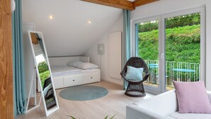 A bedroom with a bed, a white sofa, a wicker chair, and a balcony with a view of greenery.