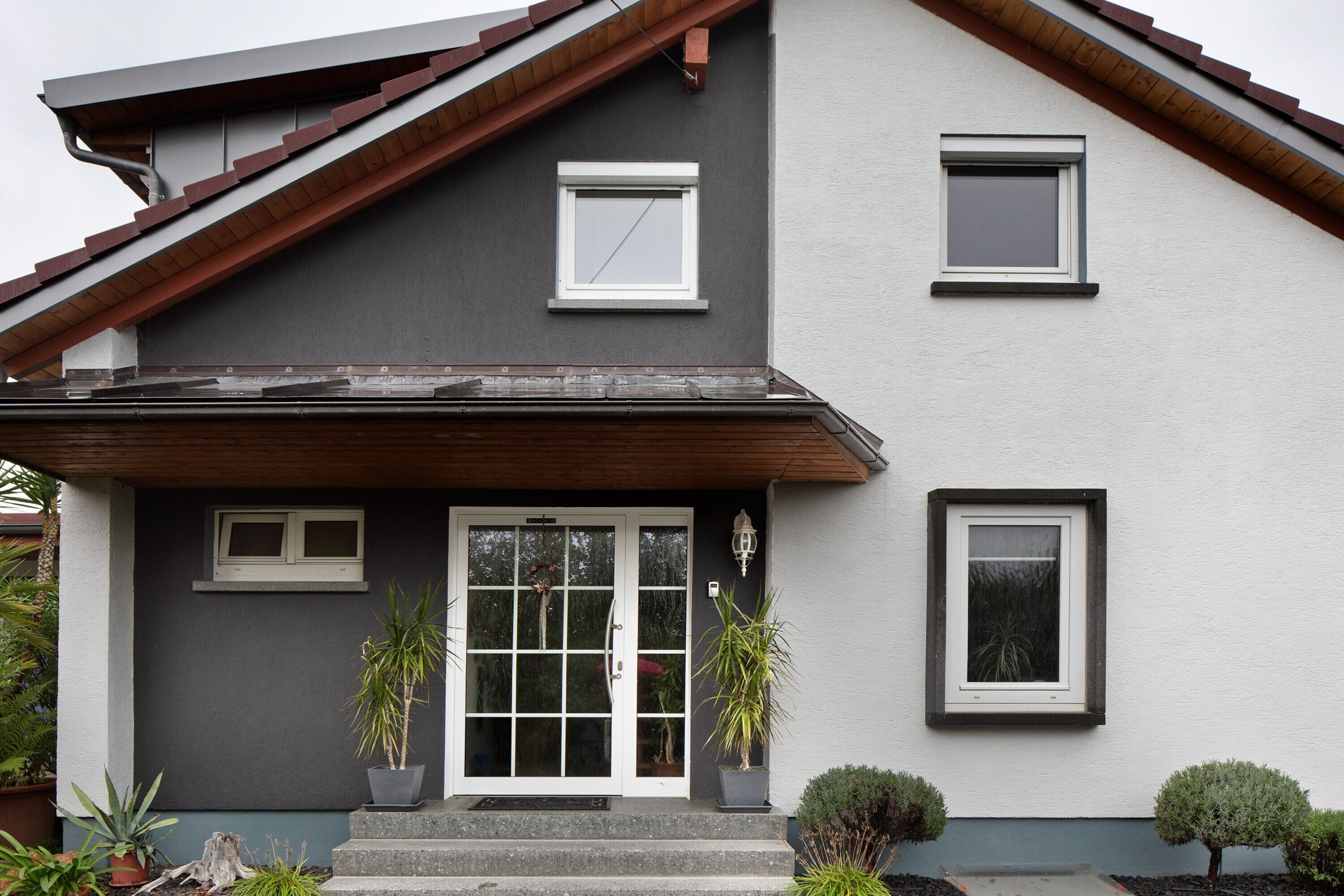 A two-story house with a grey and white exterior, a front door with glass panels, and two potted plants on the porch.
