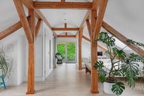 Quarto com pé-direito alto, vigas de madeira aparentes, uma grande planta monstera em vaso e vista para uma varanda com vegetação.