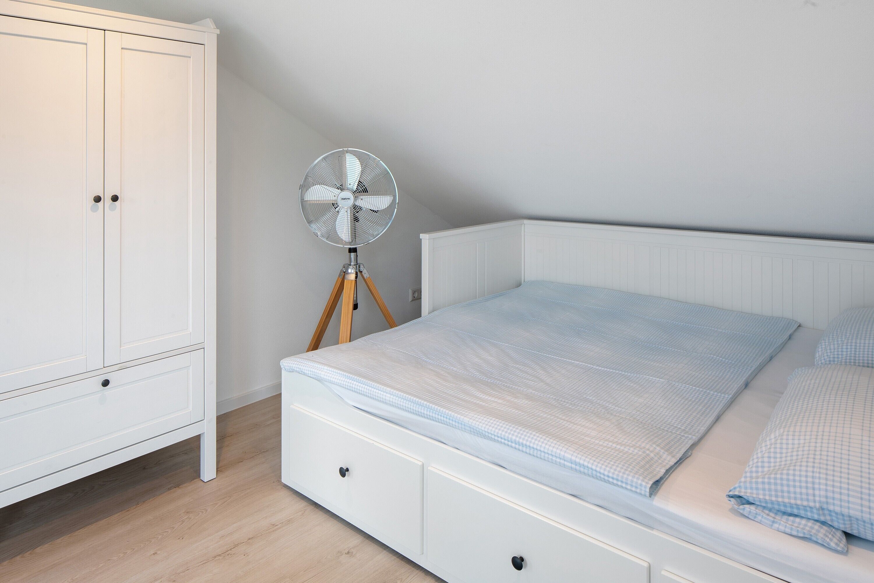 A bedroom with a white wardrobe, a bed with a blue checkered bedspread, and a standing fan.