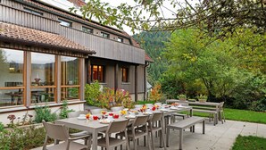 An outdoor dining area with a long table and chairs, surrounded by greenery and a building with large windows.