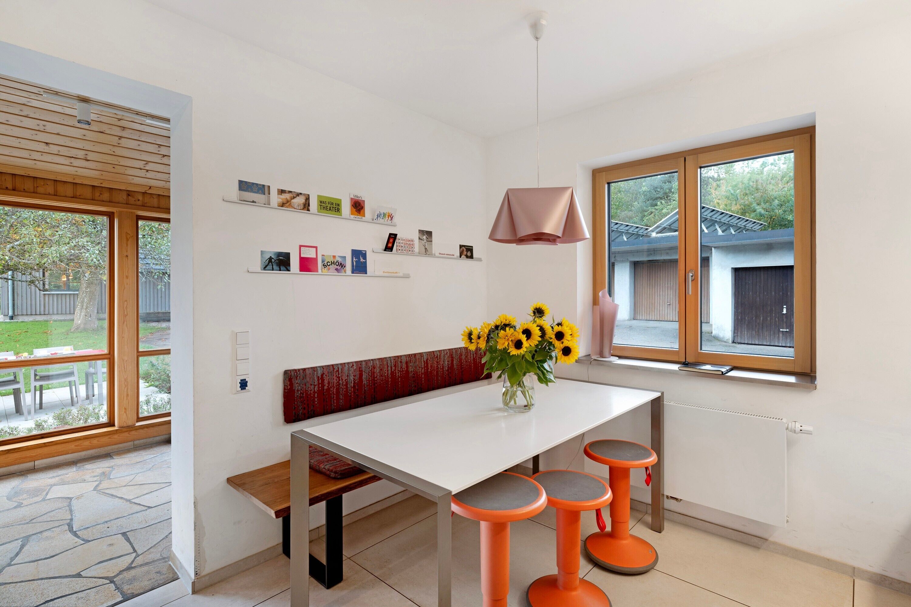 Een moderne keuken met een witte tafel, oranje krukken, een bank en een vaas met zonnebloemen.