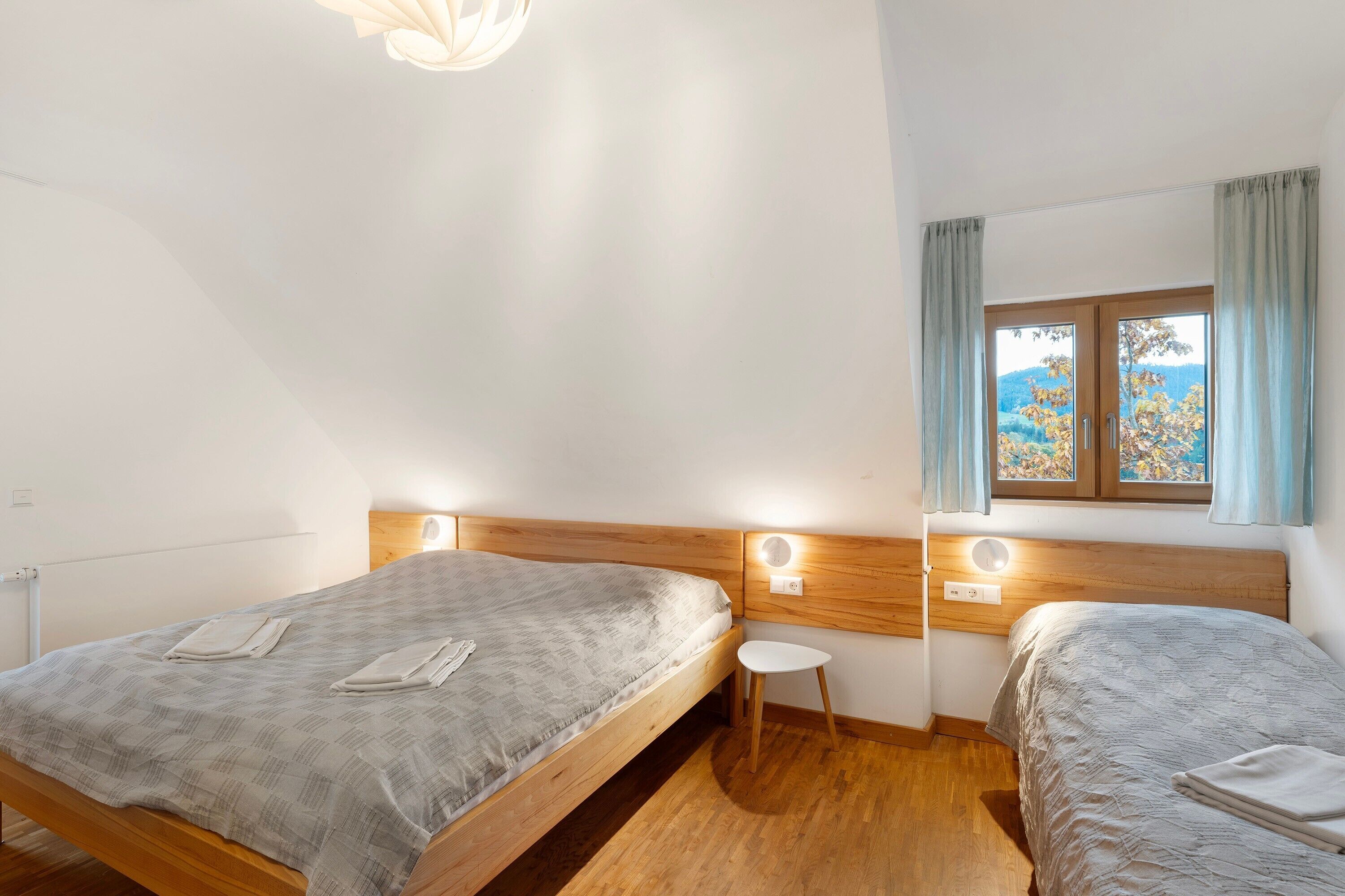 A bedroom with two beds, a small stool, a wooden headboard, a window with curtains, and a view of trees.
